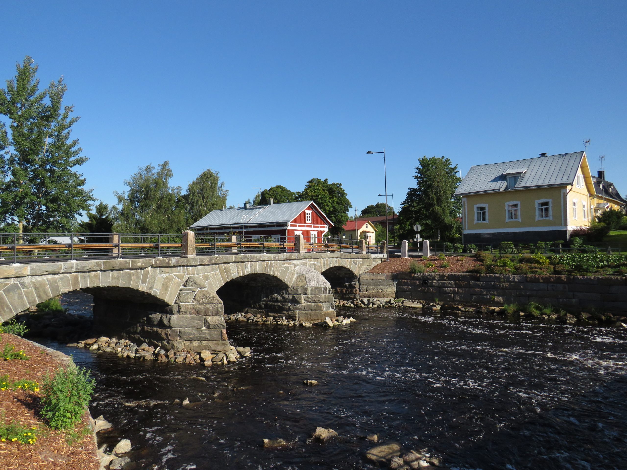Kuvassa uudistettu vanha kivisilta, joka johtaa joen toiselle puolelle. Joen toisella puolella vasemmalla punainen ja oikealla puolella keltainen puutalo.