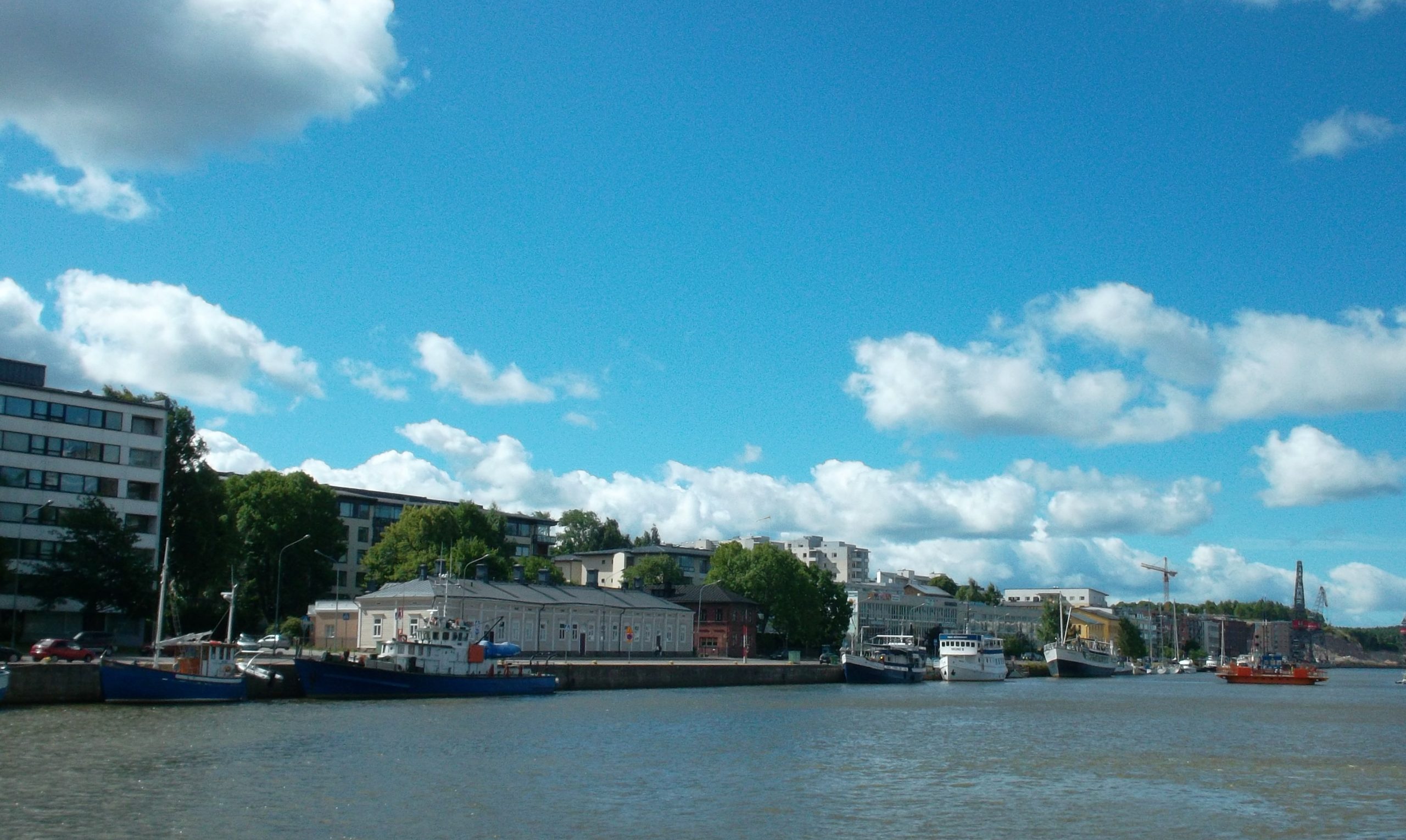 Joelta päin otetussa kuvassa näkyy erilaisia pienehköjä aluksia joessa ja rannassa eri aikakausien rakennuksia. Lähimpänä yksikerroksinen vaalea puurakennus ja seitsemänkerroksinen kerrostalo.