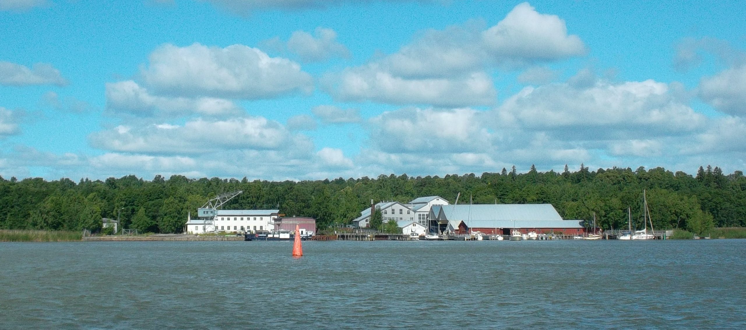 Mereltä päin otetussa kuvassa näkyy veneitä laitureissa ja useita valkoisia tai punaisia pitkänomaisia rakennuksia. Taustalla puita.