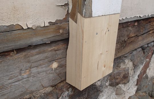 Lähikuvassa puutalon kjivijalkaa sekä hirsiarinan ja vaakalaudoituksen päällä oleva korjattu pystylauta.
