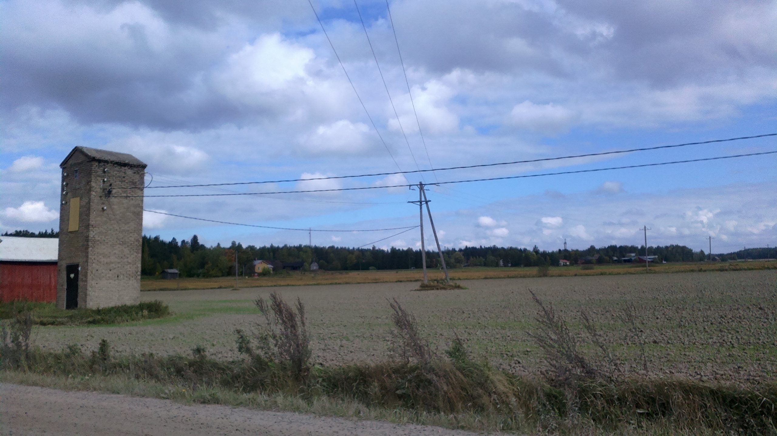 Kuvassa on pelto, jonka vasemmassa laidassa on sähkökeskus, tornimainen vaalea rakennus. Keskuksesta lähtee useita sähkökaapeleita ja sekä pellolla että pellon laidassa on useita puisia sähkötolppia.