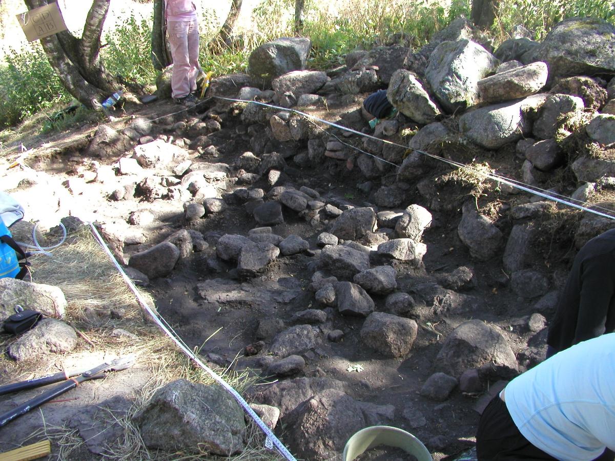 Kuvassa on suorakaiteen muotoinen kuoppa, jonka pohjalla on harmaakiviä. Kiviä on myös kuopan reunoilla, joilla näkyy myös arkeologien työvälineitä kuten ämpäreitä.