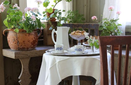 Sisäkuva puutalon kuistilta, jossa kaksi puutuolia pienen pyöreän pöydän ympärillä. Pöytä on katettu kahvihetkeä varten. Pienellä sivupöydällä on vaaleanpunaisia pelargonioita ruukuissa.