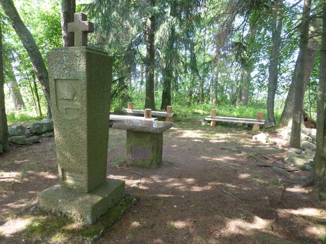 Kuvassa etualalla metsäaukealla seisova kivimonumentti, jossa risti päällä. Takana istuinpaikkoja ja toinen matala kivimonumentti.