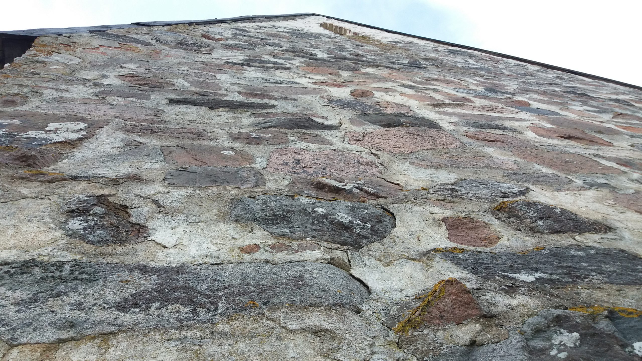 Lähikuvassa vanhan kivikirkon seinää kuvattuna seinän juurelta niin, että katonharja näkyy korkealla kuvan yläosassa.