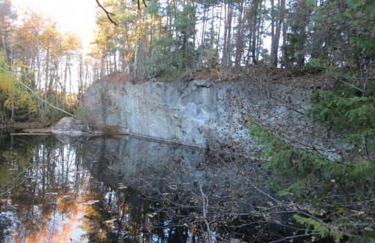 Kuvassa on keskellä tyyntä vettä, jonka takareunasta nousee ylös leikattu kallioseinämä. Kallion päällä kasvaa puita.