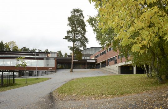 Kuvassa näkyy koulurakennus ja sen etupiha. Rakennuksen keskellä on matala pääsisäänkäynti, joka yhdistää rakennuksen kaksikerroksiset siivet toisiinsa. Vasemmanpuoleinen siipi on lasiseinäinen ja oikeanpuoleinen siipi on punatiilinen.