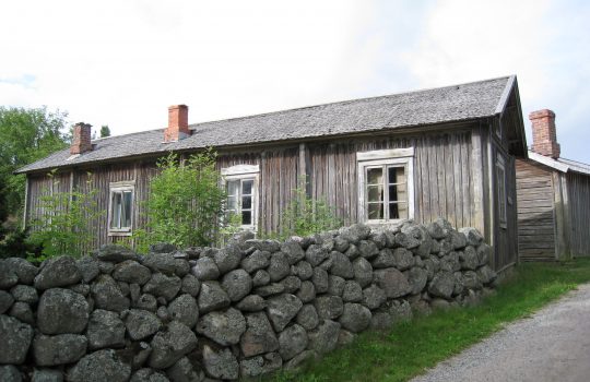 Kuvassa on harmaakivenmurikoista tehty aita, joka päättyy harmaan yksikerroksisen puutalon nurkalle. Talon kolmen ikkunan puitteet ovat valkoiset.