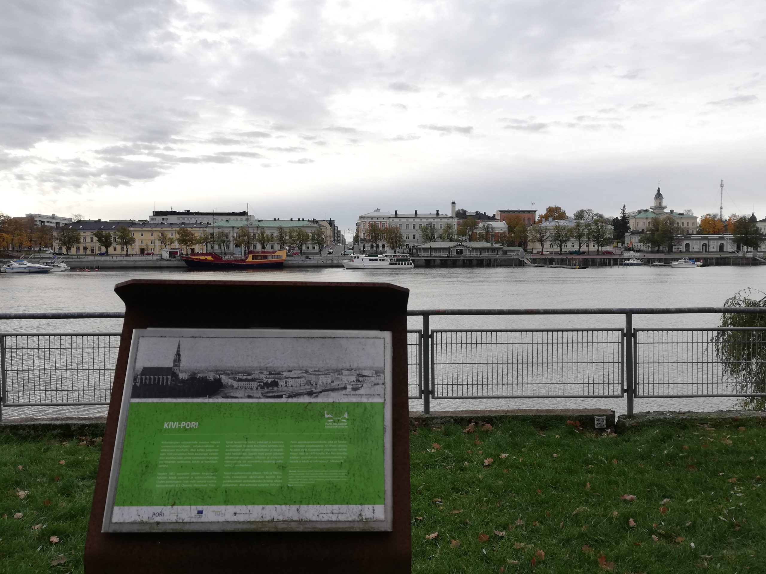 Kuvassa etualalla opaskyltti, joka kertoo Kivi-Porin historiasta. Taustalla joen toisella puolella näkyy kivirakennuksia.