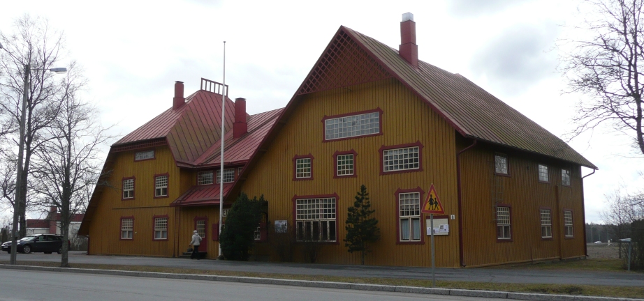 Kuvassa kellertävä puurakennus, jossa punainen peltikatto. Rakennuksen kattoa on korkea kummastakin päästä, keskellä matala. 
