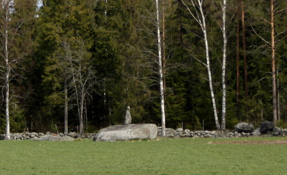 Kuvassa nurmen keskellä kivi, jonka päällä kaksi kiveä, jotka muodostavat "pään" ja "vartalon".