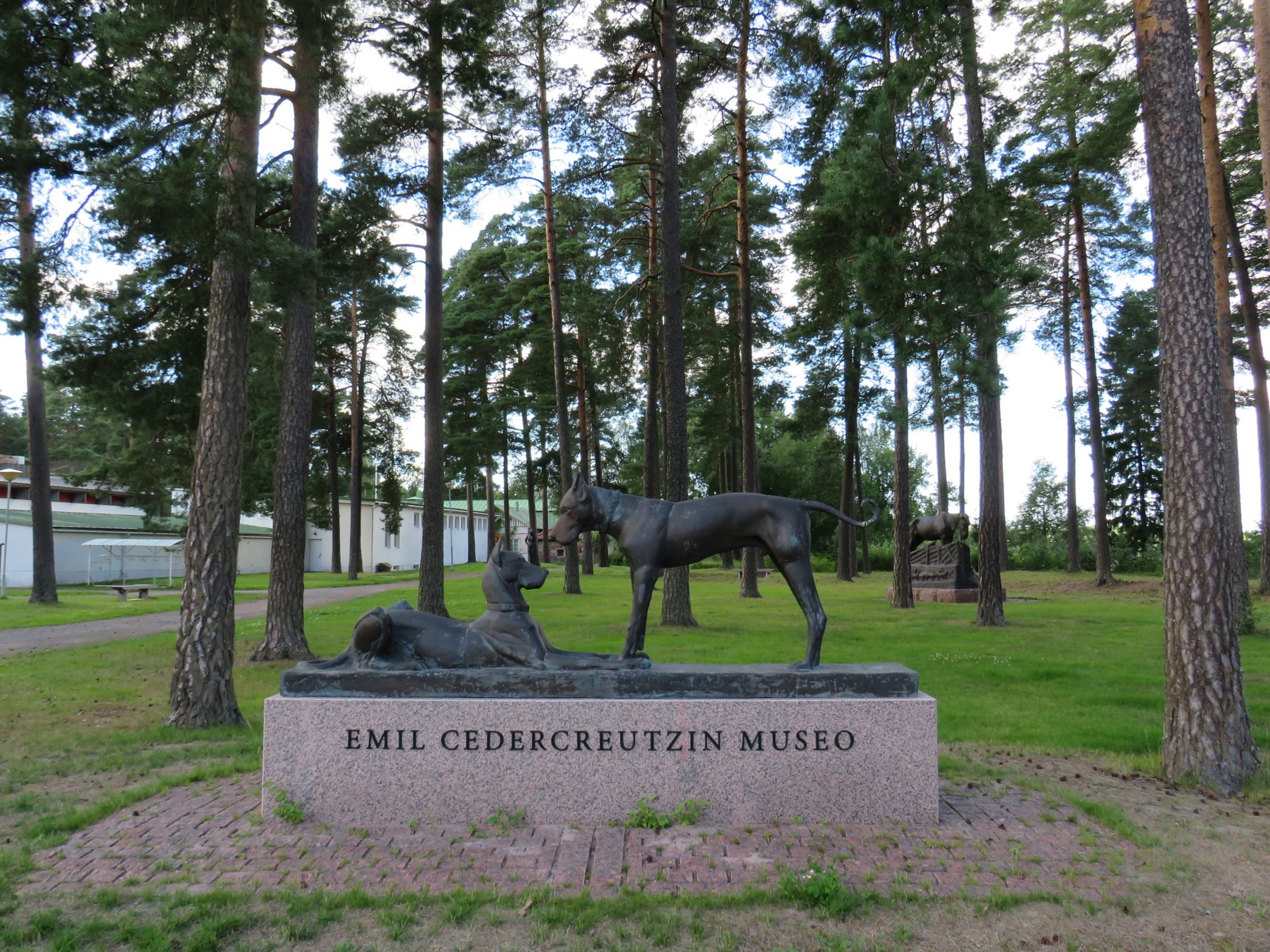Kuvassa keskellä koiria kuvastava patsas, jonka alla kivessä lukee Emil Cedercreutzin museo. Taustalla näkyy vasemmalla museo, keskellä metsää ja muita patsaita. 