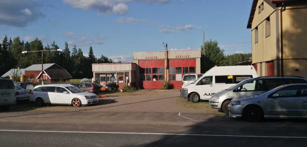Asvalttipihan molemmille reunoille parkkeerattu autoja. Pihan perällä laatikkomainen tasakattoinen rakennus, jonka etuseinä on punainen ovi.